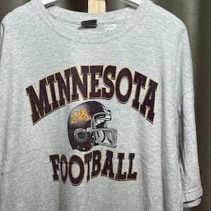 Könnte beinhalten: Graues T-Shirt mit dem Wort "MINNESOTA" in dunkelbraunen Buchstaben, das sich oben wölbt. Darunter befindet sich ein Football-Helm mit einem goldenen "M". Das Wort "FOOTBALL" ist darunter in Dunkelbraun gedruckt.
