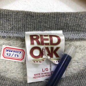 May include: Close-up of a gray sweatshirt collar with a tag that reads "RED OAK SPORTSWEAR" in maroon lettering. The tag also indicates a size "L/G" and "MADE IN HONDURAS". A small tag with the numbers "WSW01017 23/27" is also visible.