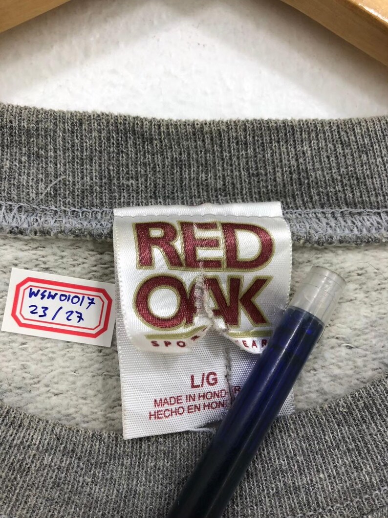 May include: Close-up of a gray sweatshirt collar with a tag that reads "RED OAK SPORTSWEAR" in maroon lettering. The tag also indicates a size "L/G" and "MADE IN HONDURAS". A small tag with the numbers "WSW01017 23/27" is also visible.