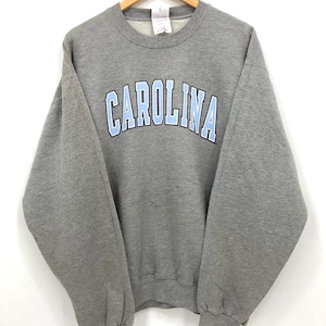 May include: A gray crewneck sweatshirt with the word "CAROLINA" arched across the chest in light blue letters. The sweatshirt is hanging on a wooden hanger against a white background. The garment appears to be made of a soft, comfortable material.