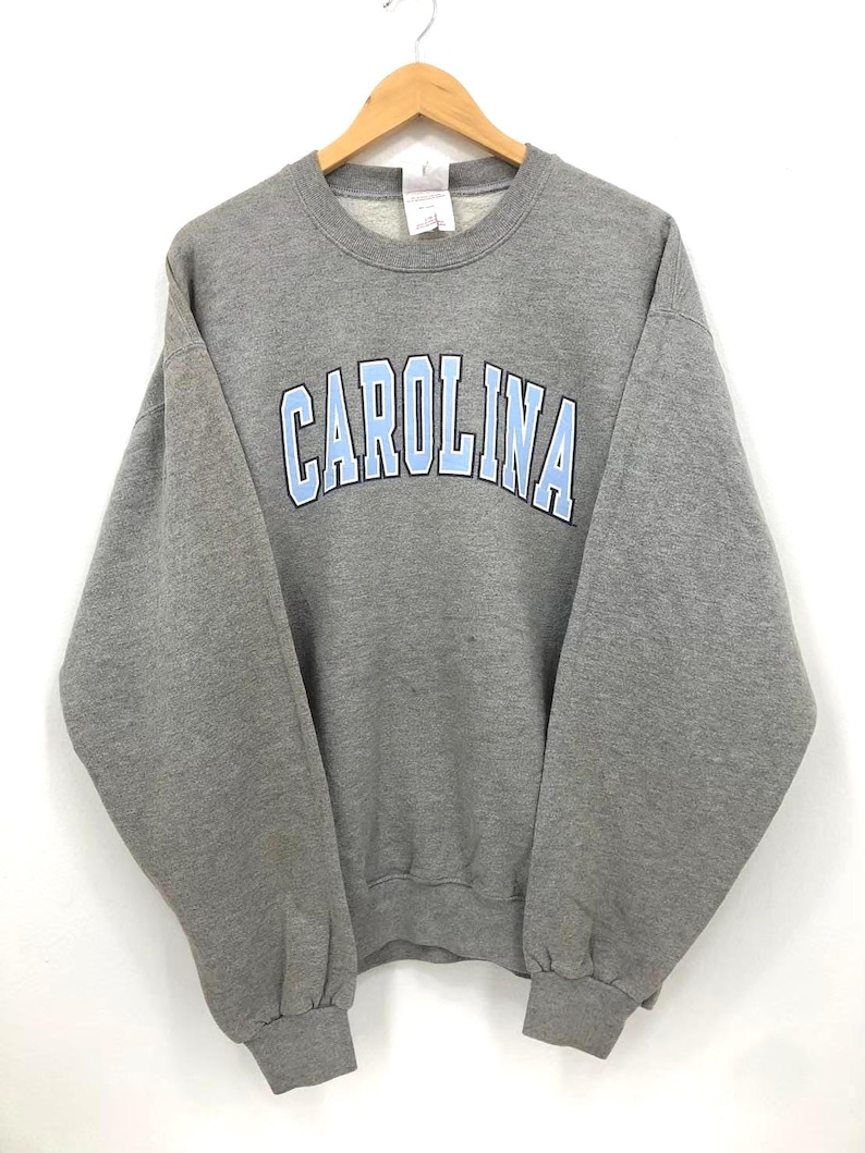 May include: A gray crewneck sweatshirt with the word "CAROLINA" arched across the chest in light blue letters. The sweatshirt is hanging on a wooden hanger against a white background. The garment appears to be made of a soft, comfortable material.