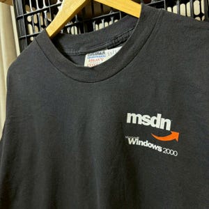 May include: A black short-sleeved t-shirt featuring the white text msdn with an orange arrow graphic and the white text Microsoft Windows 2000 below it. The shirt has a Gildan Heavyweight Cotton brand label visible at the inner neckline.