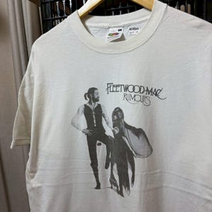 May include: Cream-coloured t-shirt with a black and white image of two figures and the words "Fleetwood Mac Rumours". The tee has a round neck and short sleeves. The label reads "Fruit of the Loom".