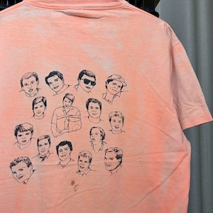 Puede incluir: Camiseta de color melocotón con una colección de retratos esbozados en blanco y negro. Los retratos son de varias personas, dispuestas en grupo en la parte trasera de la camiseta. La camiseta tiene cuello redondo y mangas cortas.