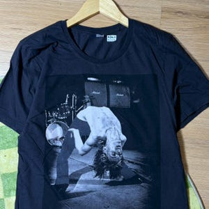 以下が含まれることがあります： ステージで演奏するミュージシャンの白黒画像がプリントされた黒いTシャツ。画像には、ドラムセットとアンプを背景に、後ろに曲がっているミュージシャンが写っています。シャツはクルーネックで半袖です。