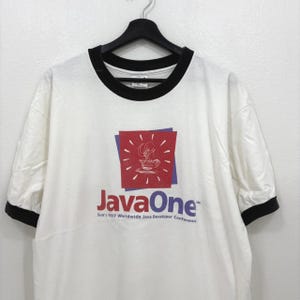 Puede incluir: Camiseta blanca con ribetes negros en el cuello y las mangas. La camiseta presenta un gráfico con un diseño de taza de café dentro de un cuadrado rojo, con el texto "JavaOne" y "Sun's 1997 Worldwide Java Developer Conference".