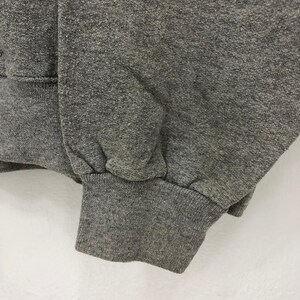 May include: Close-up of a gray sweatshirt sleeve and cuff. The fabric has a textured appearance. The cuff is ribbed and gathered at the wrist. The background is a plain white surface.