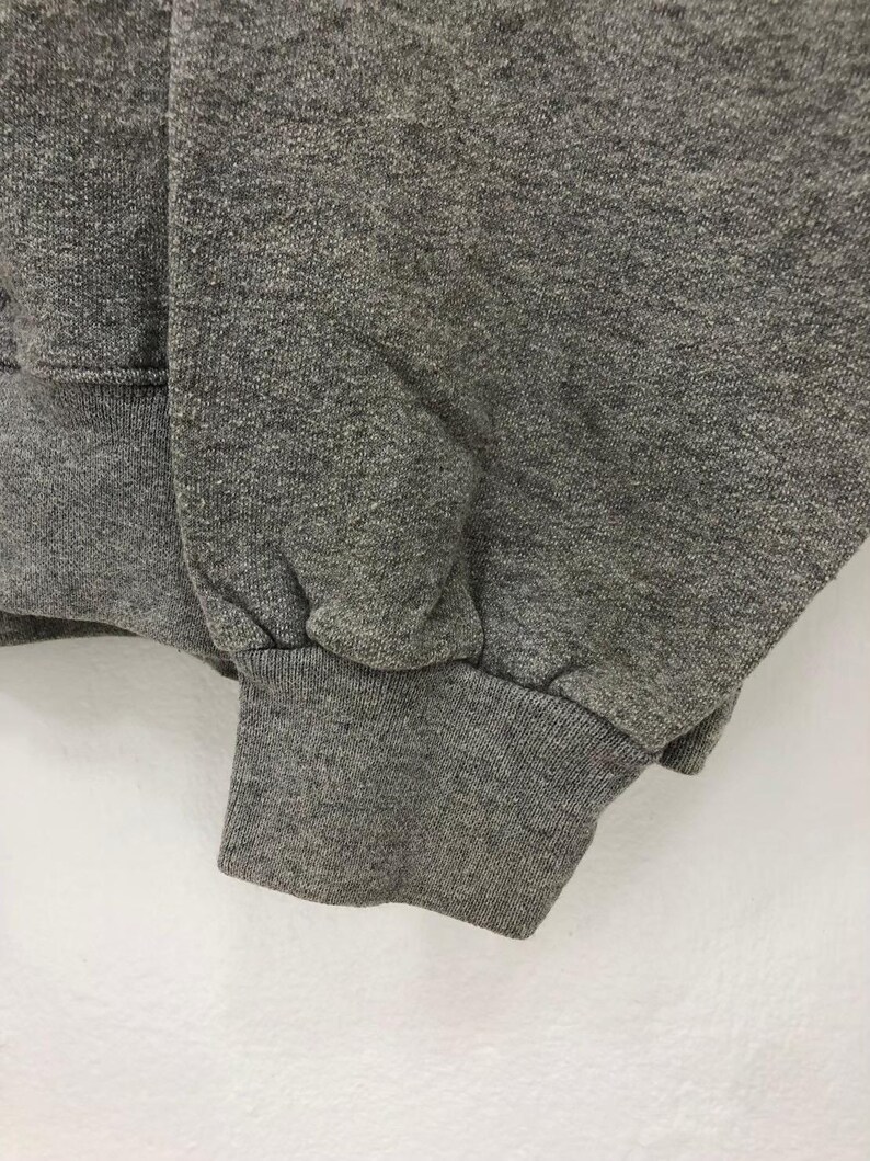 May include: Close-up of a gray sweatshirt sleeve and cuff. The fabric has a textured appearance. The cuff is ribbed and gathered at the wrist. The background is a plain white surface.