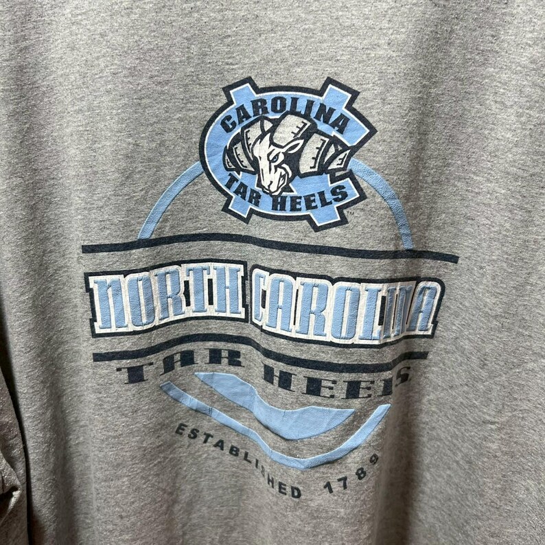 May include: A gray crew neck t-shirt featuring the words "North Carolina Tar Heels" in a light blue and white design. The shirt also includes the text "Established 1789" and a logo with a ram.