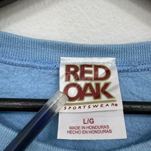 以下が含まれることがあります： 赤と白で「RED OAK SPORTSWEAR」と書かれたタグが付いた、水色のスウェットシャツのクローズアップ。タグにはサイズL/Gと「MADE IN HONDURAS HECHO EN HONDURAS」も記載されています。
