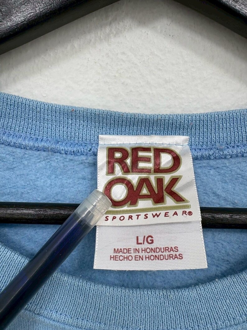 以下が含まれることがあります： 赤と白で「RED OAK SPORTSWEAR」と書かれたタグが付いた、水色のスウェットシャツのクローズアップ。タグにはサイズL/Gと「MADE IN HONDURAS HECHO EN HONDURAS」も記載されています。