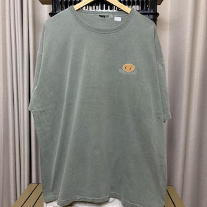 K&ouml;nnte beinhalten: Ein verwaschenes olivgr&uuml;nes T-Shirt mit kurzen &Auml;rmeln. Auf der linken Brust befindet sich ein kleines, kreisf&ouml;rmiges Logo in Orange und Gelb. Das Logo zeigt das Wort "Tradition" in geschwungener Schrift. Das T-Shirt h&auml;ngt an einem Holzb&uuml;gel.