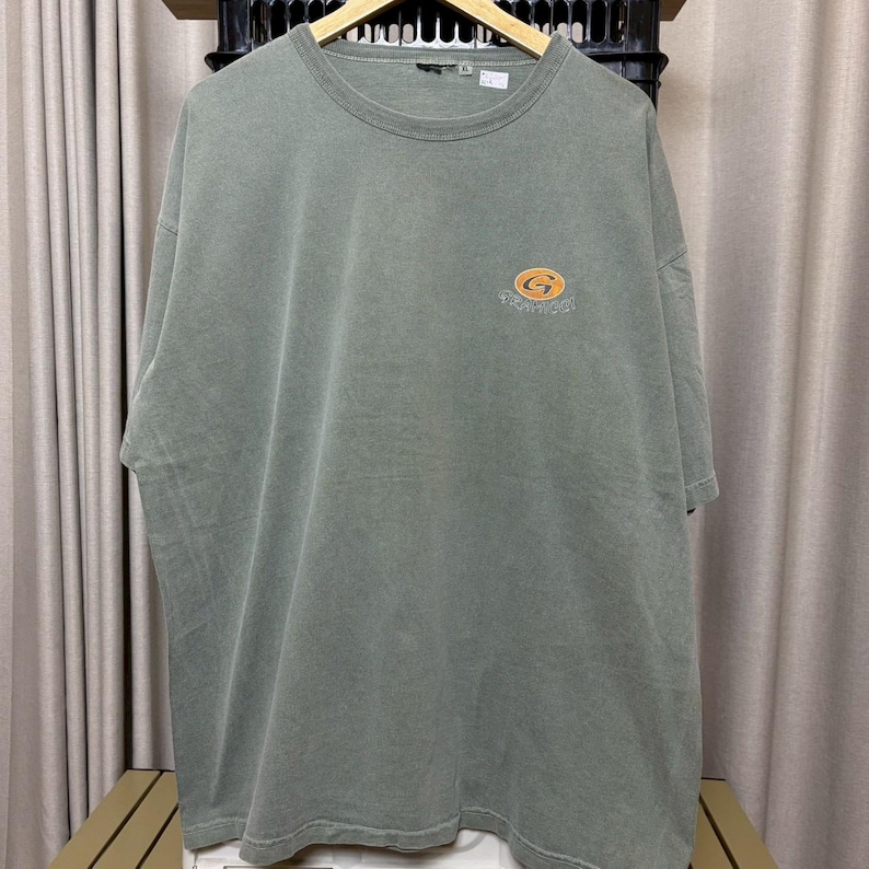 K&ouml;nnte beinhalten: Ein verwaschenes olivgr&uuml;nes T-Shirt mit kurzen &Auml;rmeln. Auf der linken Brust befindet sich ein kleines, kreisf&ouml;rmiges Logo in Orange und Gelb. Das Logo zeigt das Wort "Tradition" in geschwungener Schrift. Das T-Shirt h&auml;ngt an einem Holzb&uuml;gel.