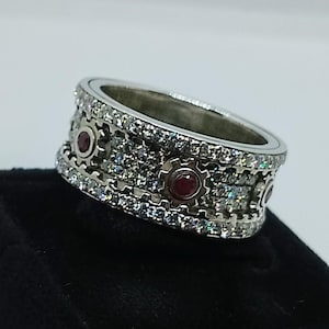 May include: A silver ring with a band featuring alternating small red gems and clear, sparkling stones. The ring is set on a black display stand, showcasing its intricate design and craftsmanship.