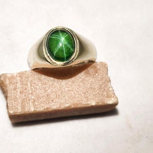 May include: A gold ring with a large, oval-shaped green gemstone. The gemstone has a star-like pattern. The ring is set on a small, rectangular, brown stone against a white background.
