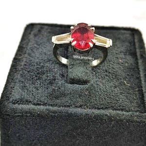 Peut inclure: Une bague en argent ornée d'une grosse pierre rouge foncé de forme ovale au centre. Deux pierres plus petites, rectangulaires et de couleur claire flanquent la pierre principale. La bague est présentée sur une surface en velours noir. Le texte "Mita jewellers" est visible.