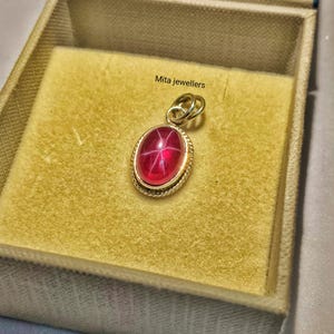 May include: A gold pendant with a large, oval, red gemstone, set in a gold frame. The gemstone has a star-like effect. The pendant is inside a beige jewelry box. The text "Mita jewellers" is visible.
