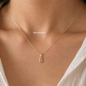 Op de afbeelding: Een delicate gouden ketting met een hanger in de vorm van een cursieve letter "C". De hanger hangt aan een fijne ketting. De ketting wordt getoond tegen een wit overhemd. De tekst "Mita jewellers" is zichtbaar.