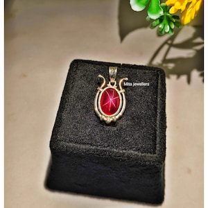 Pode incluir: Um pendente de ouro com uma pedra preciosa de rubi oval em forma de estrela. O rubi está engastado numa moldura de ouro com detalhes decorativos. O pendente é exibido numa superfície de veludo preto. O texto "Mita jewellers" é visível.