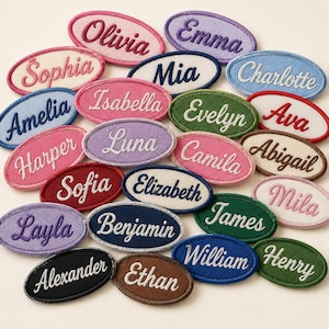 May include: A collection of oval-shaped embroidered name patches in various colors including pink, blue, green, purple, brown, and black. Each patch features a unique name stitched in a contrasting thread color, such as Olivia, Emma, Sophia, Mia, and Charlotte.