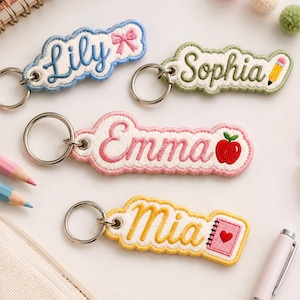 May include: Four embroidered fabric keychains with silver metal rings. The keychains feature the names Lily with a pink bow, Sophia with a yellow pencil, Emma with a red apple, and Mia with a pink notebook, each with a scalloped border.