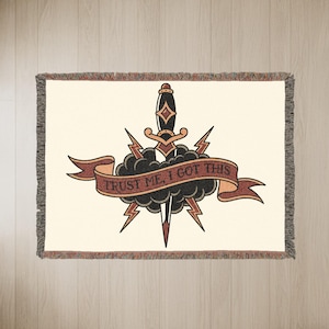 May include: Cream-colored woven throw blanket featuring a tattoo-style design. The design includes a dagger, lightning bolts, and a banner with the text "TRUST ME, I GOT THIS" in black. The blanket has a fringed edge.