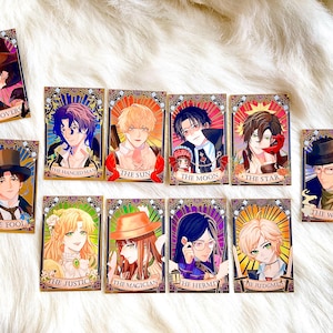 May include: A collection of tarot cards featuring anime-style illustrations. Each card has a unique character design and is labeled with titles like "The Lovers," "The Fool," and "The Sun." The cards are set against a soft, white background.