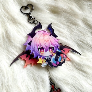 May include: A colorful anime-style keychain featuring a character with pink and purple hair, large eyes, and bat-like wings. The keychain has a black heart-shaped clasp and a silver chain. The character is holding a small object. The wings are red and black.