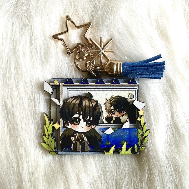May include: A gold-colored star-shaped keychain with a blue tassel. The keychain features a colorful anime-style illustration of two characters in a window, surrounded by green leaves. The background is white.