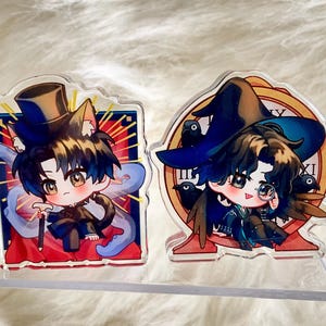 May include: Two acrylic charms featuring anime-style characters. One character wears a top hat and cat ears, holding a cane. The other wears a witch hat and has crows. Both have large eyes and are set against colorful backgrounds.