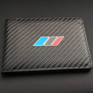 Może przedstawiać: Czarny, składany portfel z wzorem splotu z włókna węglowego. Portfel posiada kolorowy emblemat z niebieskimi, szarymi i czerwonymi akcentami. Portfel jest prostokątny i wydaje się być wykonany z trwałego materiału.