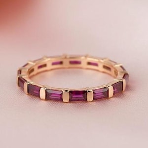 Op de afbeelding: Een dunne gouden ring met een doorlopende rij diep magenta edelstenen in baguette-slijpvorm, gezet in een kanaalzetting. De ring heeft een gepolijste metalen afwerking en een uniforme cirkelvorm, met een reeks rechthoekige stenen.