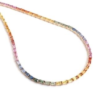 Puede incluir: Un delicado collar con un degradado de piedras preciosas de colores. Las piedras preciosas transitan del rosa al naranja, amarillo, verde, azul y morado. El collar está engastado en un metal dorado, creando una joya vibrante y elegante.