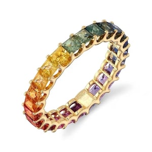 May include: A gold ring featuring a rainbow of square-cut gemstones. The gemstones transition from red to orange, yellow, green, and blue. The ring is set in a gold band, creating a vibrant and colorful piece of jewelry.