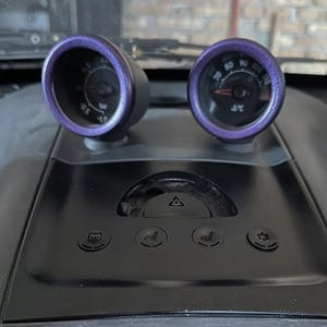 May include: Dashboard detail featuring two gauges with purple trim. The left gauge displays pressure in bar, while the right gauge shows temperature in degrees Fahrenheit. The dashboard is black with various control buttons.