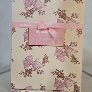 May include: A rectangular box with a floral and bow pattern in pink and white. The box is wrapped with a pink ribbon and a tag that reads "LOVESHACKFANCY" and "STANLEY". The box is likely a gift box or packaging for a product.