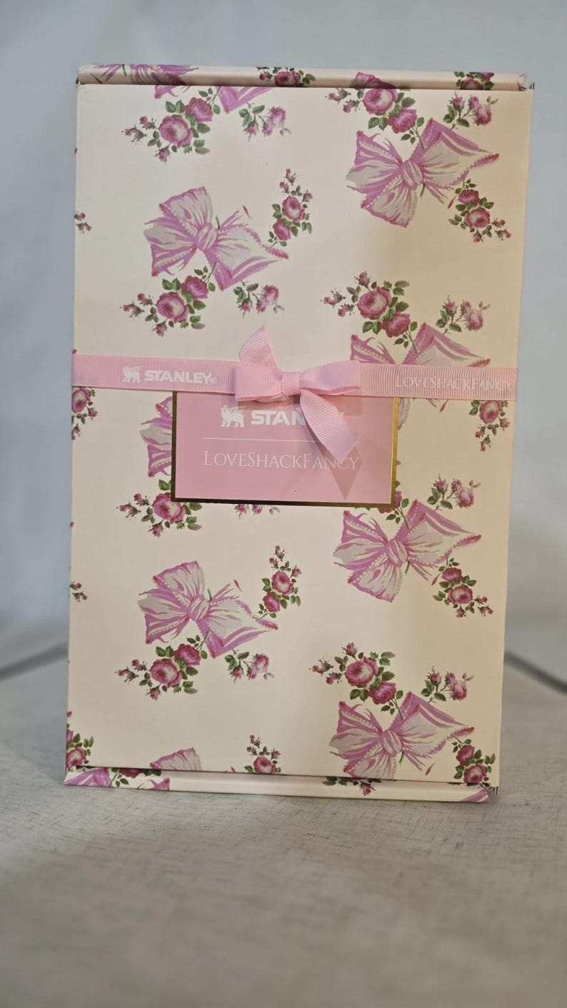 May include: A rectangular box with a floral and bow pattern in pink and white. The box is wrapped with a pink ribbon and a tag that reads "LOVESHACKFANCY" and "STANLEY". The box is likely a gift box or packaging for a product.