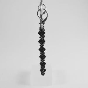 Puede incluir: Un solo pendiente de cuentas negras con un cierre plateado. El pendiente presenta un dise&ntilde;o lineal de peque&ntilde;as cuentas redondas dispuestas en un patr&oacute;n floral. El pendiente se muestra sobre un fondo blanco.