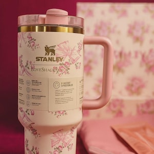 May include: A white Stanley LoveShackFancy tumbler with a pink floral and bow pattern. The tumbler has a pink handle, lid, and accents. The text "STANLEY" and "LOVESHACK FAN" are visible on the tumbler.
