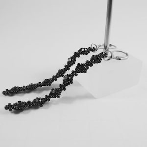 Puede incluir: Pendientes colgantes negros con cuentas. Los pendientes presentan un delicado dise&ntilde;o floral con peque&ntilde;as cuentas redondas. Cuelgan de ganchos plateados. Los pendientes se exhiben sobre una superficie blanca.