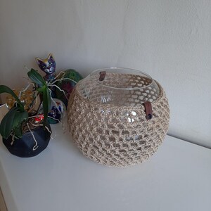 May include: A clear glass bowl with a woven beige jute cover and brown leather accents. The bowl is round and sits on a white surface. A small potted plant and decorative cat figurine are in the background.