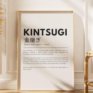 May include: A framed print with the word "KINTSUGI" in large black letters. Below, Japanese characters and the definition of Kintsugi, the art of repairing broken pottery with gold. A vase with dried plants and a bamboo side table are also in the image.