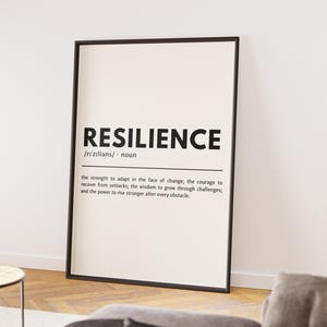May include: A framed print with the word "RESILIENCE" in bold black letters. Below, the definition of resilience is provided in smaller black text on a white background. The frame is black, and the print is displayed against a white wall.