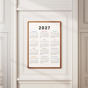 May include: A wall calendar for the year 2027 displayed in a wooden frame. The calendar features a white background with black text for the months and dates. Each month name is printed in a different color, organized in a grid of twelve.