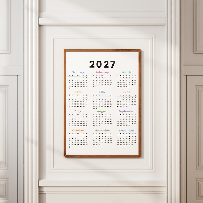 May include: A wall calendar for the year 2027 displayed in a wooden frame. The calendar features a white background with black text for the months and dates. Each month name is printed in a different color, organized in a grid of twelve.