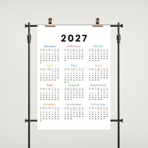 May include: A 2027 wall calendar printed on a white rectangular sheet, suspended by two metallic clips from a black horizontal rod. The calendar displays all twelve months in a grid format with black text and colorful month headers.