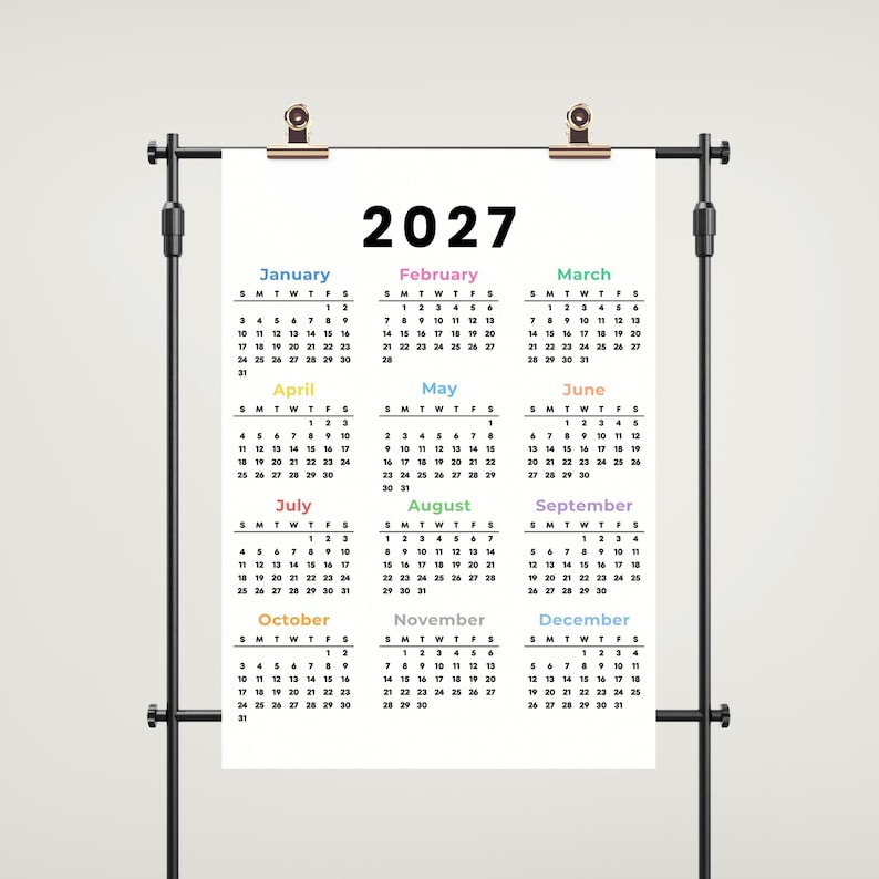 May include: A 2027 wall calendar printed on a white rectangular sheet, suspended by two metallic clips from a black horizontal rod. The calendar displays all twelve months in a grid format with black text and colorful month headers.