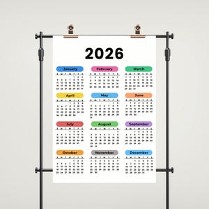 May include: A 2026 calendar displayed on a black metal stand. Each month is color-coded, with the year in bold black text at the top. The calendar is clipped to the stand with two gold clips.