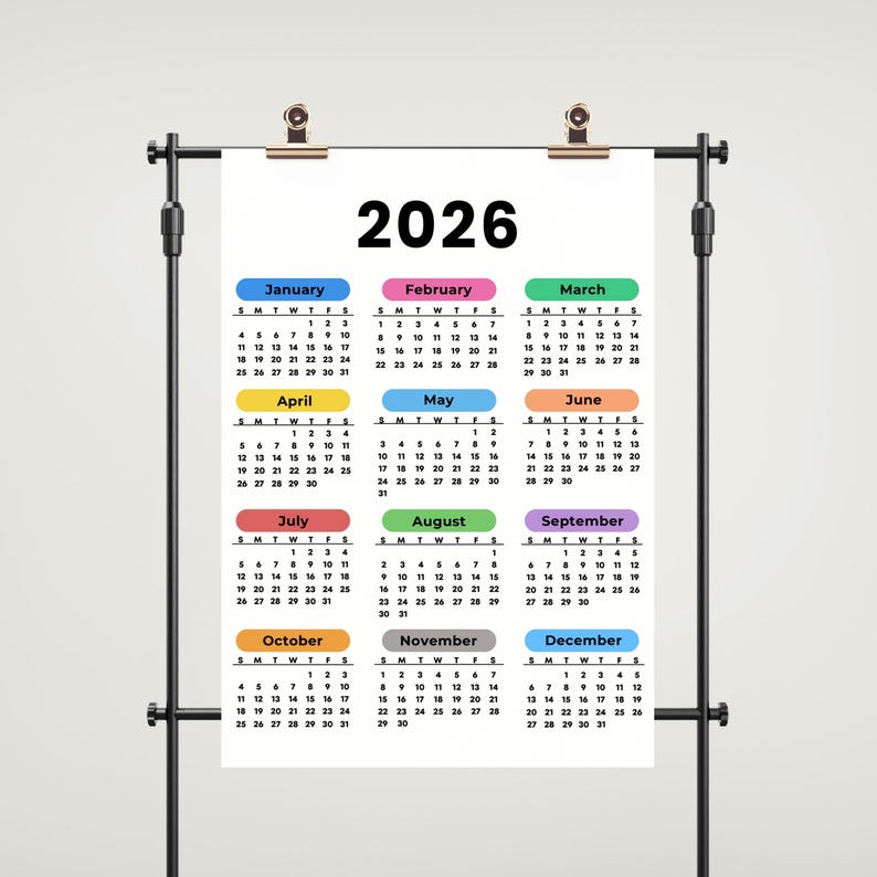 May include: A 2026 calendar displayed on a black metal stand. Each month is color-coded, with the year in bold black text at the top. The calendar is clipped to the stand with two gold clips.