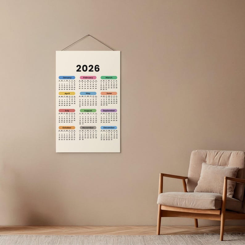 May include: A 2026 wall calendar with a cream background. The calendar has a simple design with the year at the top and the months labeled in different colors. The calendar is hanging on a wall.